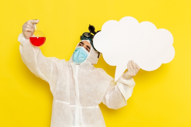 Vue avant de l'homme travailleur scientifique en tenue de protection spéciale tenant le ballon avec une solution rouge et grand panneau blanc sur un bureau jaune clair