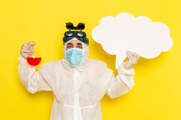 Vue avant de l'homme travailleur scientifique en tenue de protection spéciale tenant le ballon avec une solution rouge grand panneau blanc sur le bureau jaune