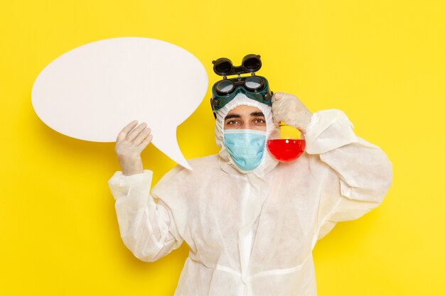 Vue avant de l'homme travailleur scientifique en tenue de protection spéciale tenant le ballon avec une solution rouge et panneau blanc sur un bureau jaune