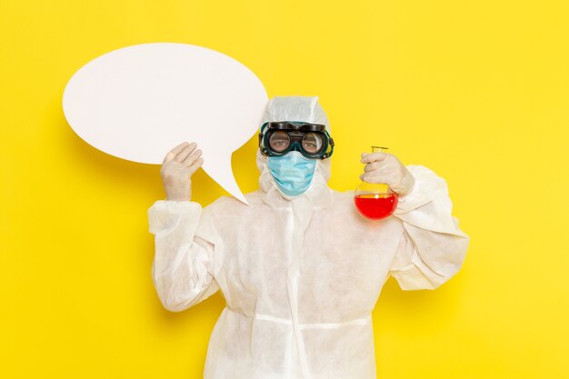 Vue avant de l'homme travailleur scientifique en tenue de protection spéciale tenant le ballon avec une solution rouge et panneau blanc sur un bureau jaune