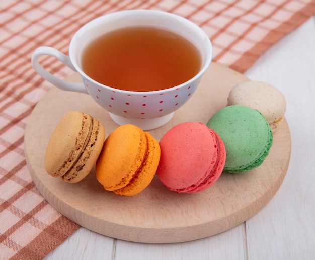 Vue avant des macarons multicolores avec une tasse de thé sur un support sur une serviette à carreaux marron