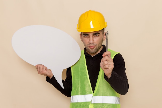 Vue avant mâle constructeur en casque jaune tenant une pancarte blanche et un outil sur fond crème