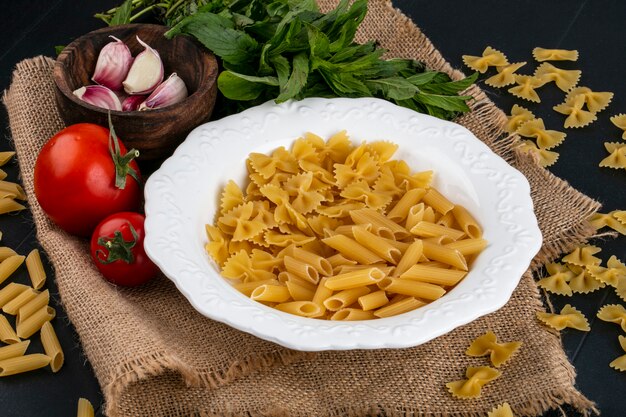 Vue avant des pâtes crues dans une assiette avec des tomates ail et un bouquet de menthe sur une serviette beige