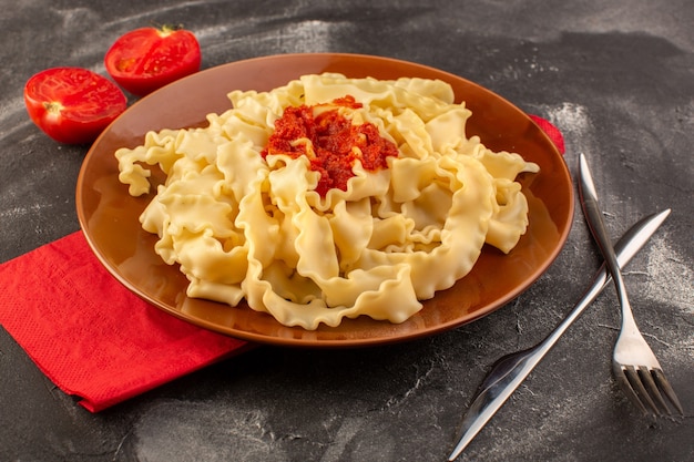 Une vue avant de pâtes italiennes cuites avec des couverts de sauce tomate et tomates à l'intérieur de la plaque sur la surface grise