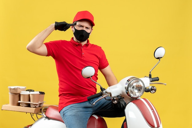 Vue avant de la pensée émotionnelle jeune adulte portant un chemisier rouge et des gants de chapeau dans un masque médical délivrant l'ordre assis sur un scooter sur fond jaune