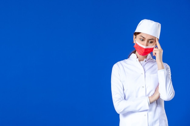 Vue avant de penser jeune infirmière en costume médical avec masque de protection rouge sur mur bleu