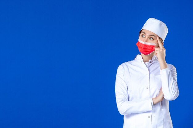 Vue avant de penser jeune infirmière en costume médical avec masque de protection rouge sur mur bleu