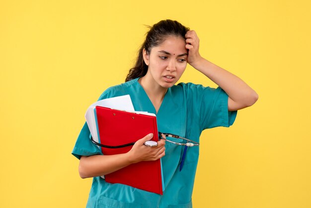 Vue avant réfléchie femme médecin avec stéthoscope tenant des dossiers dans sa main debout sur fond jaune