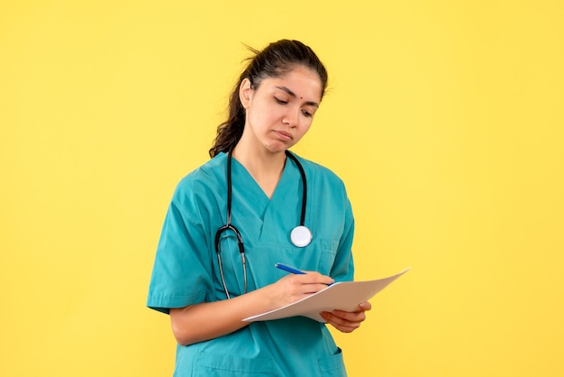 Vue avant réfléchie jolie femme médecin écrit quelque chose sur papier sur fond jaune