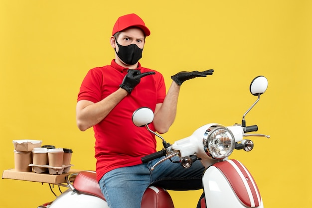 Vue avant de se demander jeune adulte portant un chemisier rouge et des gants de chapeau dans un masque médical délivrant l'ordre assis sur un scooter