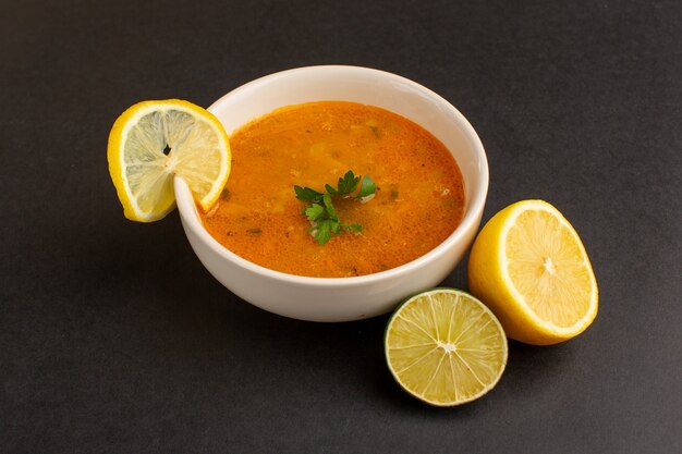 Vue avant de la soupe aux légumes savoureuse à l'intérieur de la plaque avec du citron sur un bureau sombre.