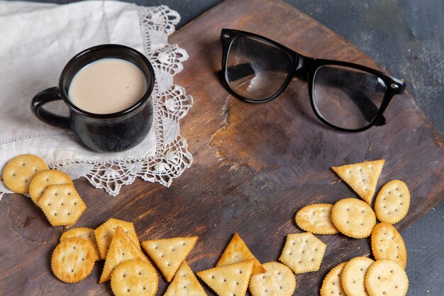 Vue avant de la tasse de lait avec des lunettes de soleil et des craquelins sur le bureau en bois et surface grise