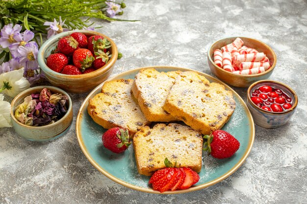 Vue avant des tranches de gâteau délicieux avec des fraises et des bonbons sur une surface légère de tartes au gâteau sucré