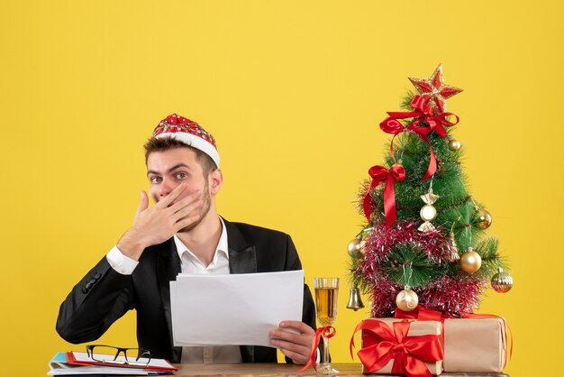 Vue avant travailleur masculin assis derrière son lieu de travail tenant des documents sur le bureau jaune nouvel an travail couleur de bureau Noël