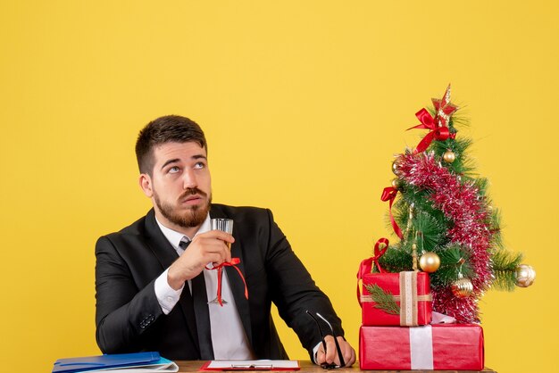 Vue avant travailleur masculin derrière sa table avec des cadeaux et arbre de Noël sur jaune