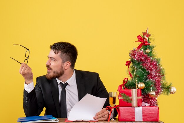 Vue avant travailleur masculin derrière sa table avec des cadeaux et arbre de Noël sur jaune