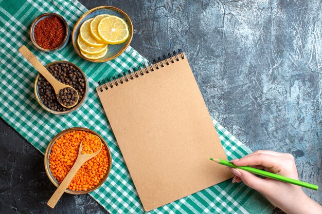 Vue ci-dessus de l'arrière-plan du dîner avec différentes épices, pois jaune et main tenant un stylo sur un cahier à spirale sur une table sombre