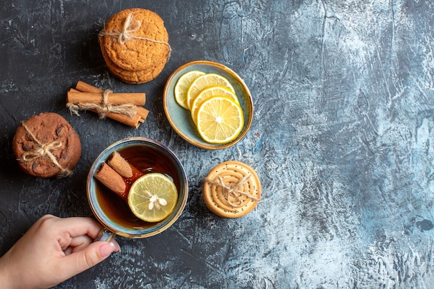 Vue ci-dessus de citrons frais et main tenant une tasse de thé noir avec des biscuits empilés à la cannelle sur fond sombre