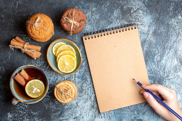 Vue ci-dessus de citrons frais et d'une tasse de thé noir à la cannelle divers biscuits empilés et main tenant un stylo cahier à spirale sur fond sombre