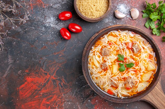 Vue ci-dessus d'une délicieuse soupe de nouilles au poulet sur une planche à découper en bois, une bouteille d'huile et une tomate à l'ail vert sur une table sombre