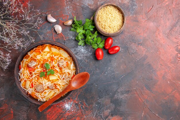 Vue ci-dessus d'une délicieuse soupe de nouilles avec du poulet et des pâtes non cuites dans un petit bol marron et une cuillère de tomates à l'ail et de légumes verts sur fond sombre