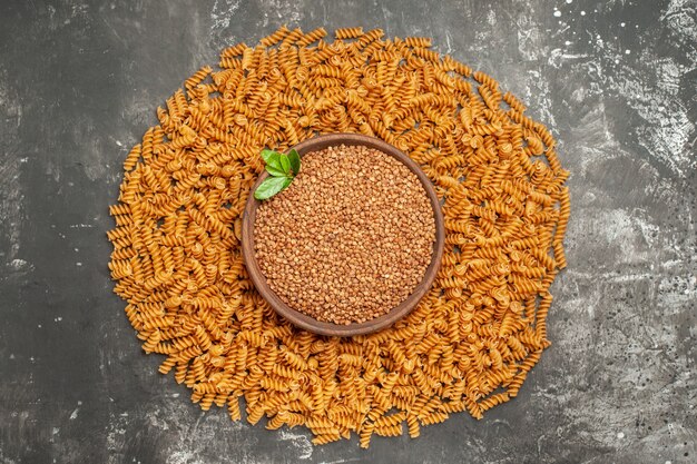Vue ci-dessus du sarrasin dans un bol marron au milieu d'un cadre circulaire bordé de pâtes italiennes crues sur fond gris
