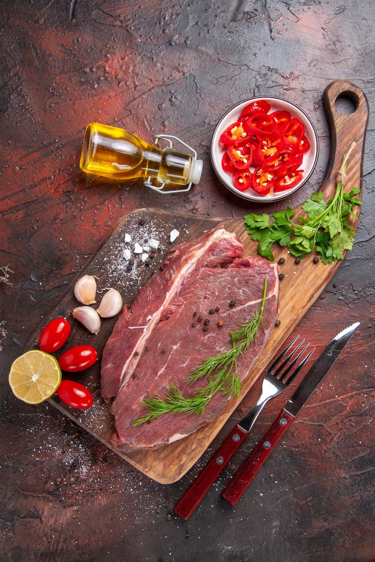 Photo gratuit vue ci-dessus de la viande rouge sur une planche à découper en bois et de l'ail vert citron haché poivre tomate bouteille d'huile tombée sur fond sombre