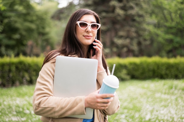 Vue côté, de, femme lunettes soleil, tenue, ordinateur portable, et, boisson, dehors
