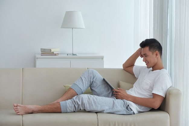 Vue de côté d'un homme asiatique assis confortablement sur le canapé et en regardant une vidéo sur son pavé numérique