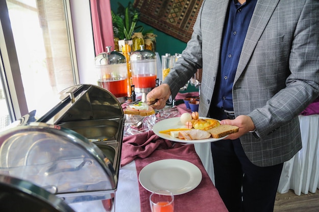 Vue de côté, l'homme pose le petit-déjeuner des saucisses aux œufs pain pain grillé et fromage sur une assiette du buffet ouvert