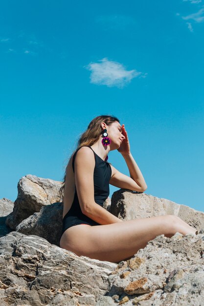 Vue de côté de la jeune femme en maillot de bain noir assis sur un rocher