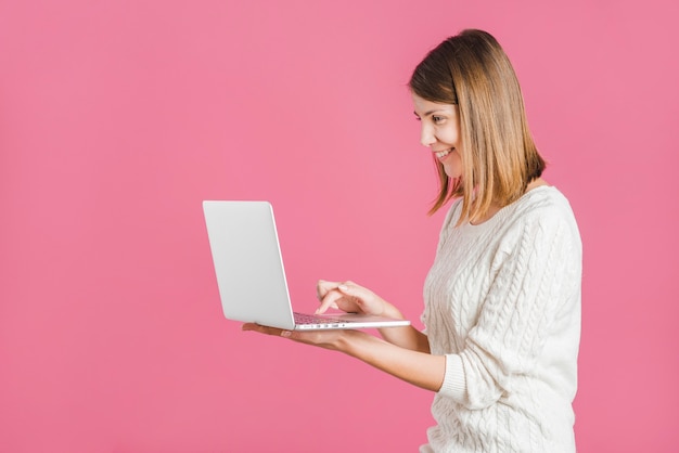Vue de côté d&#39;une jeune femme souriante travaillant sur un ordinateur portable sur fond rose