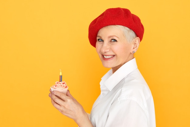 Vue de côté de la magnifique femme d'âge moyen sortie avec des cheveux teints courts sous chapeau rouge faisant souhait tenant un petit gâteau d'anniversaire avec une bougie
