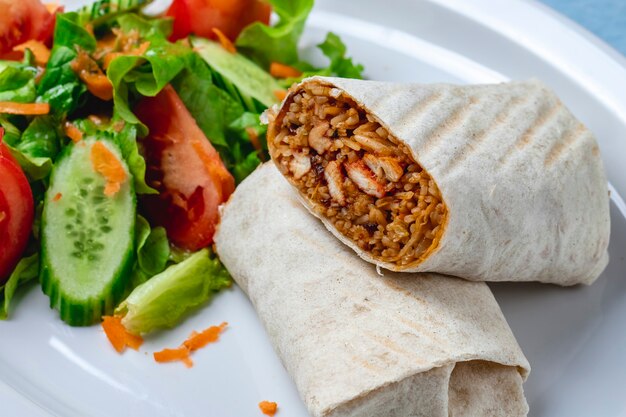 Vue de côté poulet burrito filet de poulet grillé avec du riz enveloppé dans une tortilla tomate concombre fraîche carotte et laitue sur une plaque