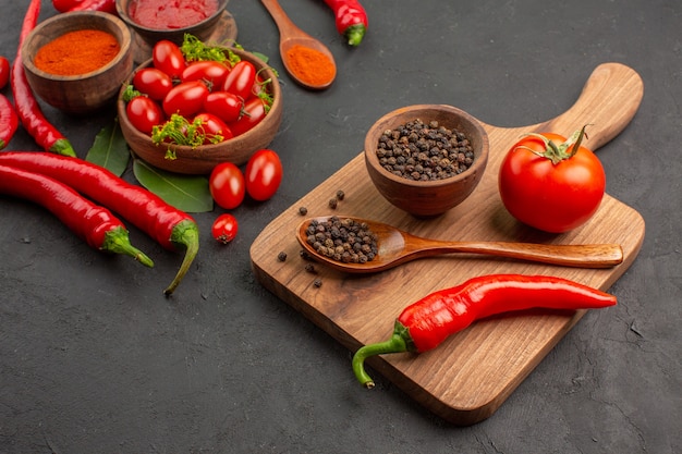 Vue de dessous un bol de tomates cerises piments rouges chauds feuilles de laurier et un bol de poivre noir une cuillère en bois un poivron rouge sur la planche à découper sur fond noir