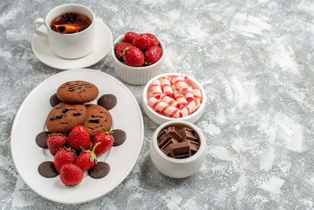 Vue de dessous cookies fraises et chocolats ronds sur la plaque ovale bols de bonbons fraises chocolats thé anis cannelle sur le côté gauche de la table gris-blanc