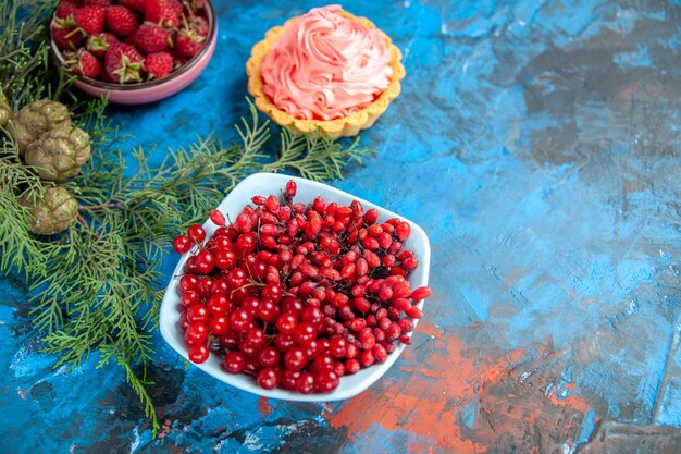 Photo gratuite vue de dessous framboises fraîches groseilles et épine-vinettes dans des bols branche de pin et cônes petite tarte sur table bleue espace libre