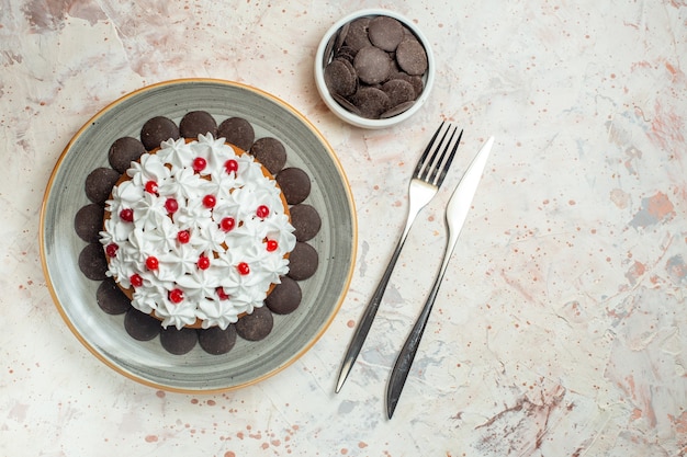 Vue de dessous gâteau avec crème pâtissière sur plaque ovale chocolat dans un bol fourchette et couteau de table sur table beige