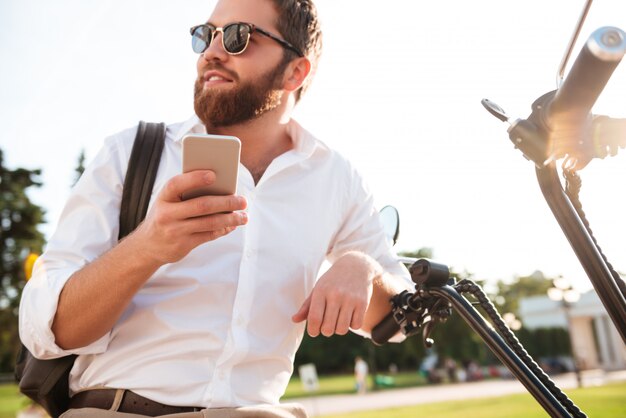 Vue de dessous d'un homme barbu souriant à lunettes de soleil assis sur une moto moderne à l'extérieur et tenant un smartphone tout en regardant ailleurs