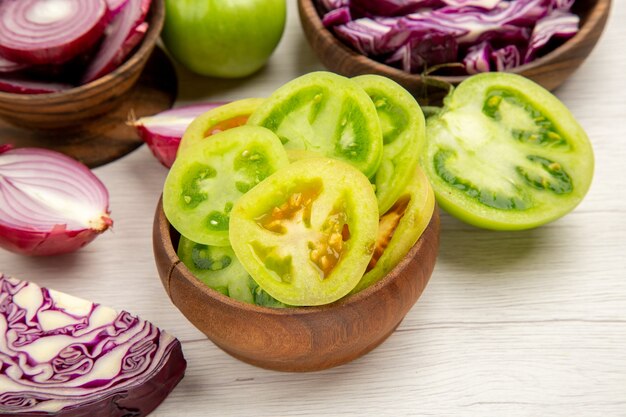 Vue de dessous légumes frais tomates vertes choux rouges oignons coupés dans des bols sur table en bois blanc