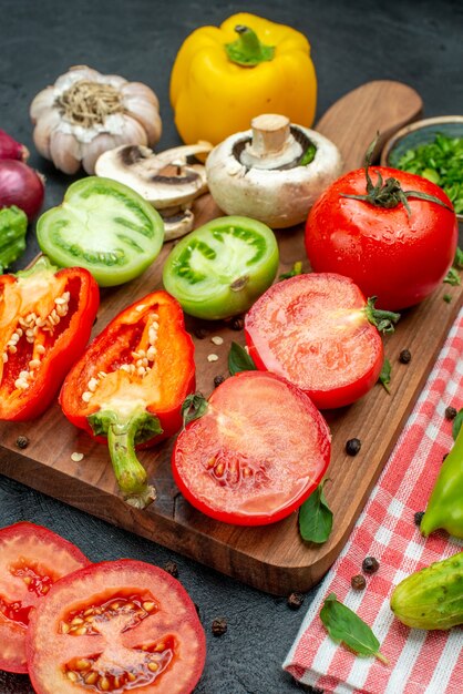 Vue de dessous légumes tomates vertes et rouges poivron jaune sur planche à découper verts dans un bol couteau concombres sur nappe rouge sur table noire