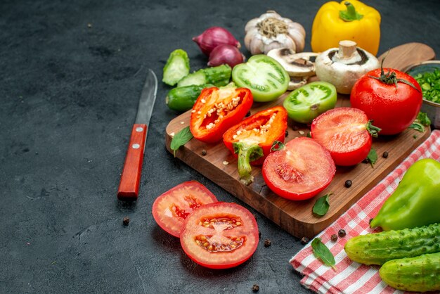 Vue de dessous légumes tomates vertes et rouges poivrons sur planche à découper verts dans un bol couteau concombres sur nappe rouge sur tableau noir