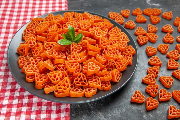 Vue de dessous des pâtes italiennes rouges en forme de coeur sur une plaque ovale noire sur une serviette de cuisine des pâtes au coeur éparpillées sur une table sombre