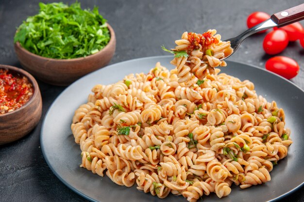 Vue de dessous des pâtes rotini sur une assiette sur une fourchette, des tomates cerises, de la sauce tomate et des légumes verts hachés dans des bols sur une surface isolée sombre