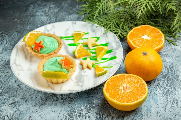 Vue de dessous de petites tartes avec de la crème pâtissière verte et une tranche de citron sur une assiette d'oranges coupées sur une surface sombre