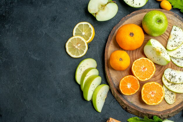 Vue de dessous des pommes et des mandarines coupées sur une planche ronde rustique Cannelle coupée des citrons cumcuat et d'autres trucs autour de la planche sur un espace libre de table sombre