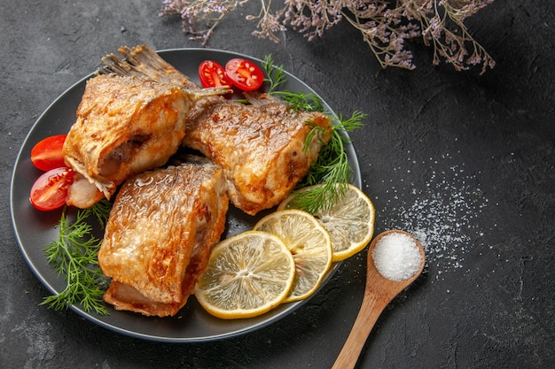 Vue de dessous de savoureux poissons frits tranches de citron coupées de tomates cerises sur une assiette branche de fleurs séchées cuillère en bois sur tableau noir