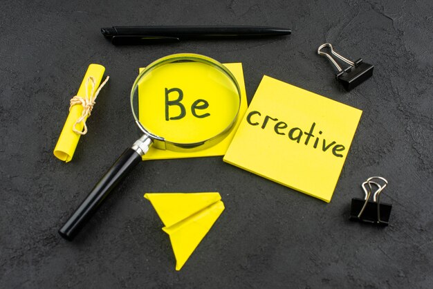 Vue de dessous soyez créatif écrit sur une note collante jaune, un stylo à pinces pour reliure lupa sur fond sombre