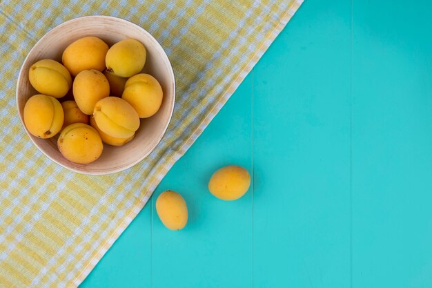 Vue de dessus des abricots dans un bol sur une serviette à carreaux jaune sur une surface bleue