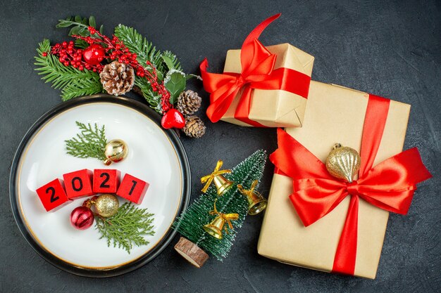 Vue de dessus des accessoires de décoration numéros sur une assiette branches de sapin conifère et coffrets cadeaux avec ruban rouge en forme d'arc sur fond sombre